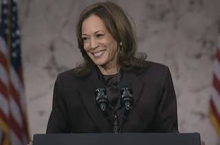 A person in a professional suit speaks at a podium with microphones, flags in the background