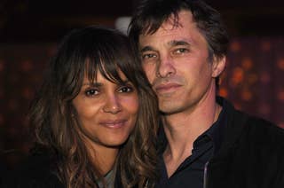 Halle Berry and Olivier Martinez stand closely together, smiling softly at the camera. Halle is wearing a casual outfit, and Olivier is in a dark jacket