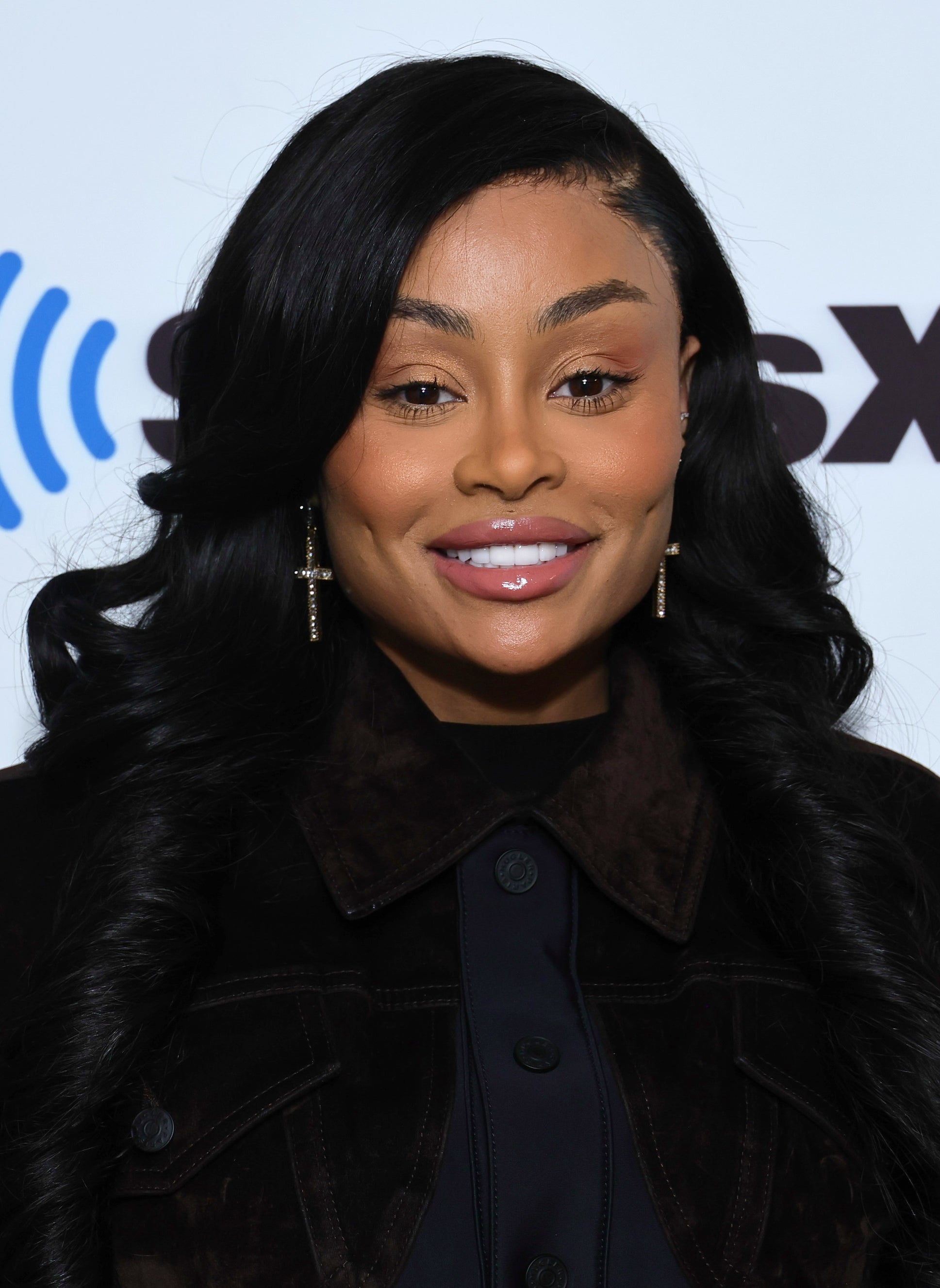 Blac Chyna poses in a dark buttoned-up outfit in front of a SiriusXM backdrop, smiling for the camera