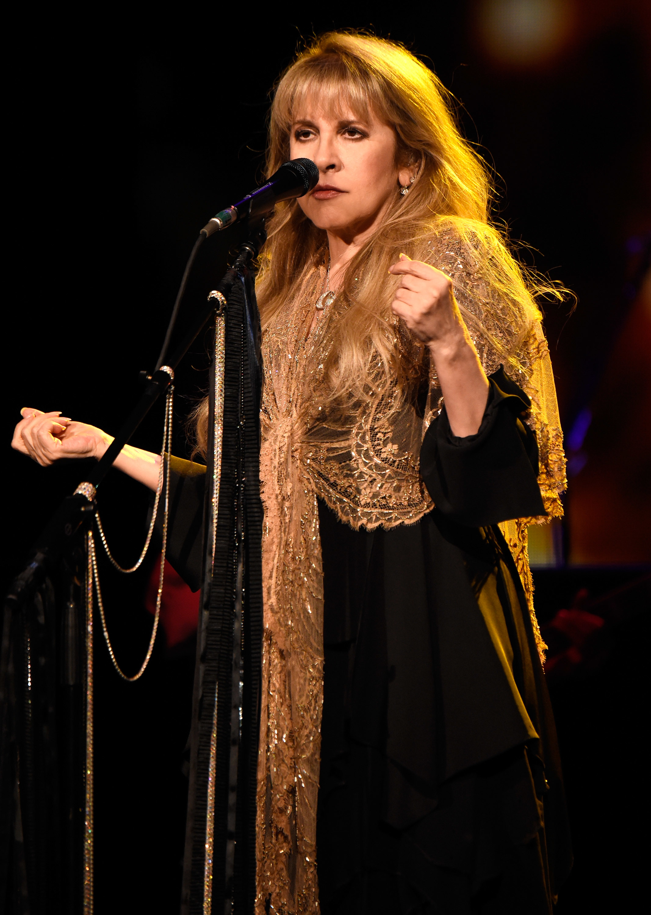 Stevie Nicks in a flowing outfit and lace shawl sings into a microphone on stage