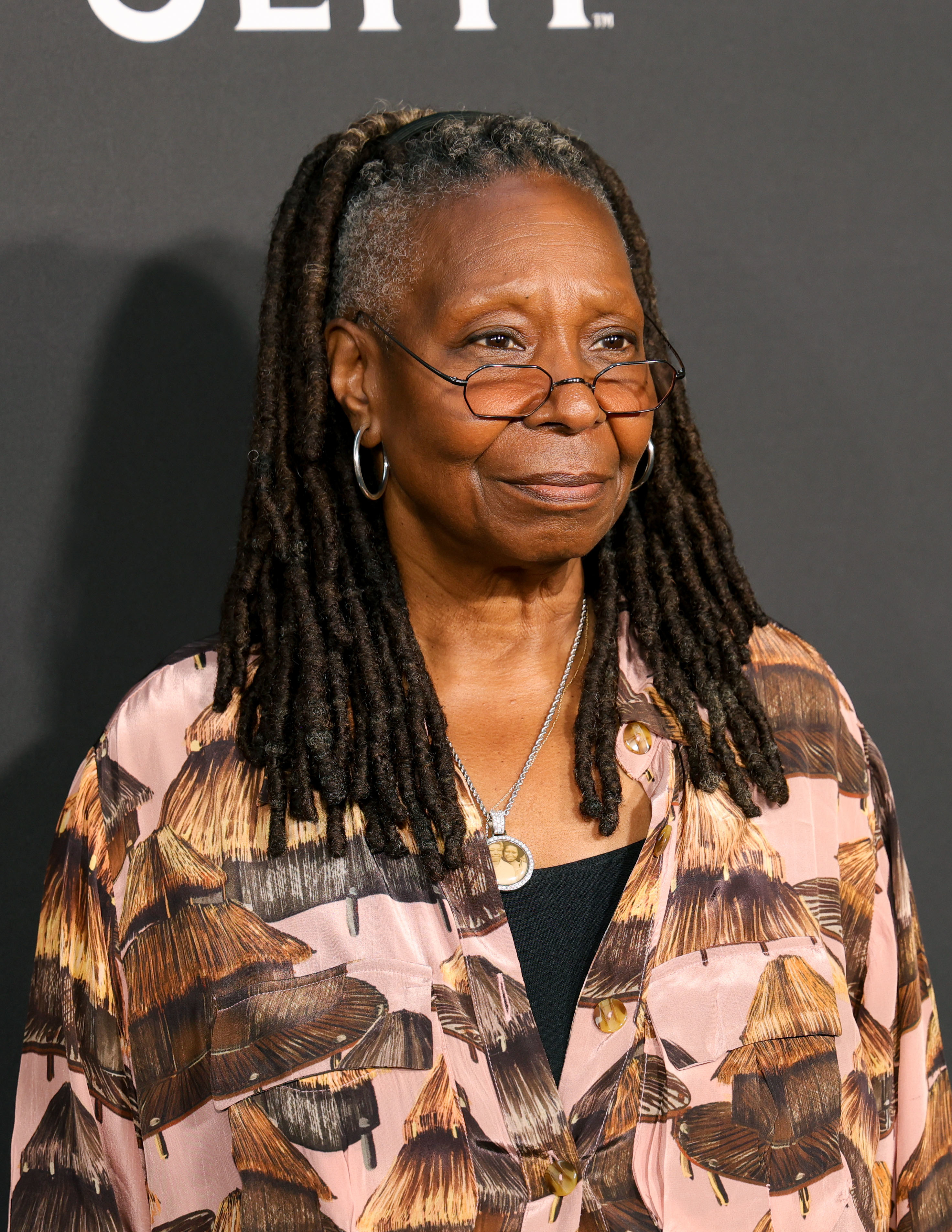 Whoopi Goldberg wearing a patterned top and glasses, standing in front of a plain background