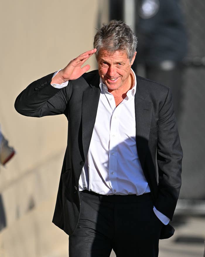 Hugh Grant smiles as he walks outside wearing a blazer and slacks