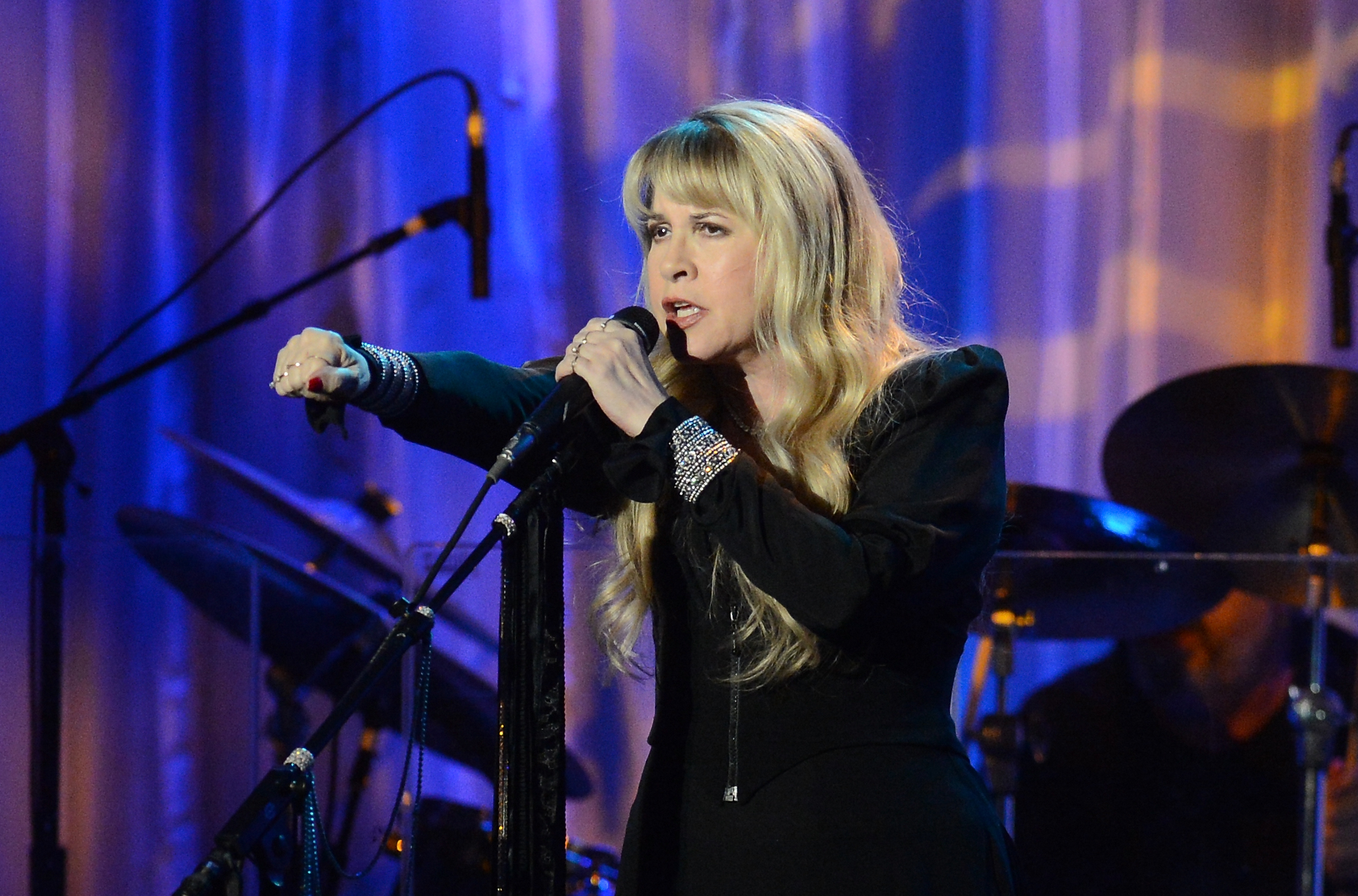 Stevie Nicks in elegant attire sings passionately on stage, holding a microphone