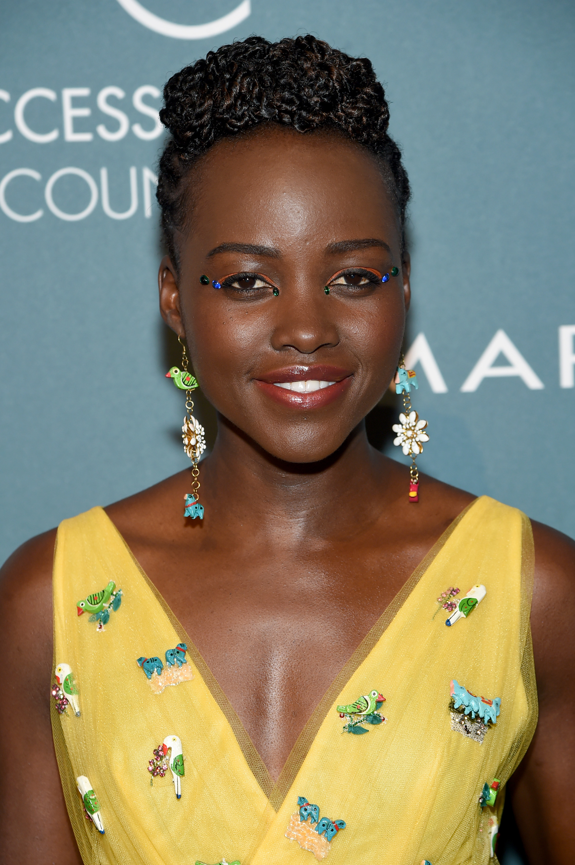 A closeup of Lupita smiling at an event wearing a sleeveless dress decorated with birds