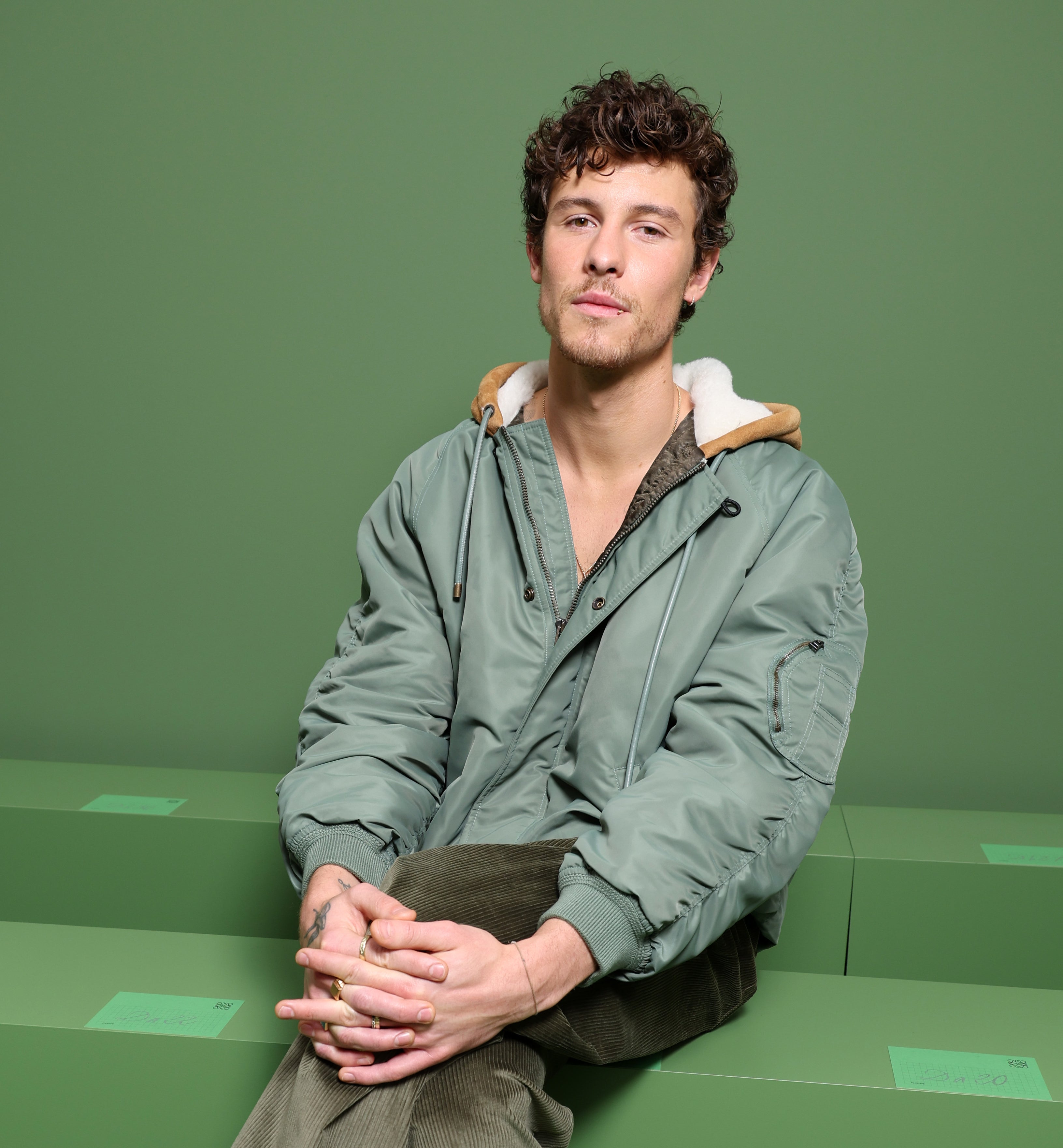 Shawn Mendes sits casually on a green bench, wearing a relaxed green bomber jacket, green pants, and black boots