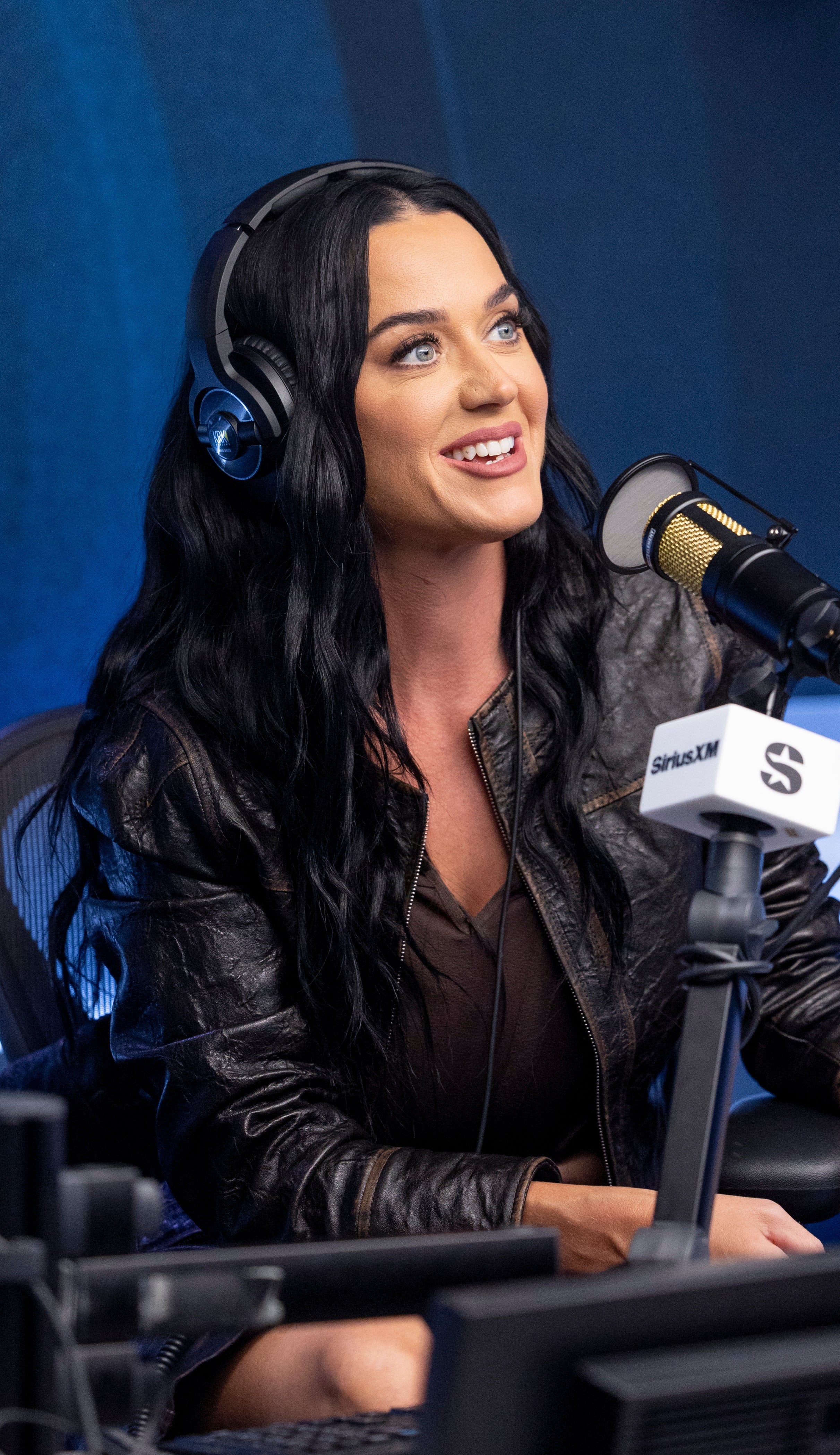 Katy Perry in a radio studio, smiling and speaking into a microphone, wearing headphones and a leather jacket