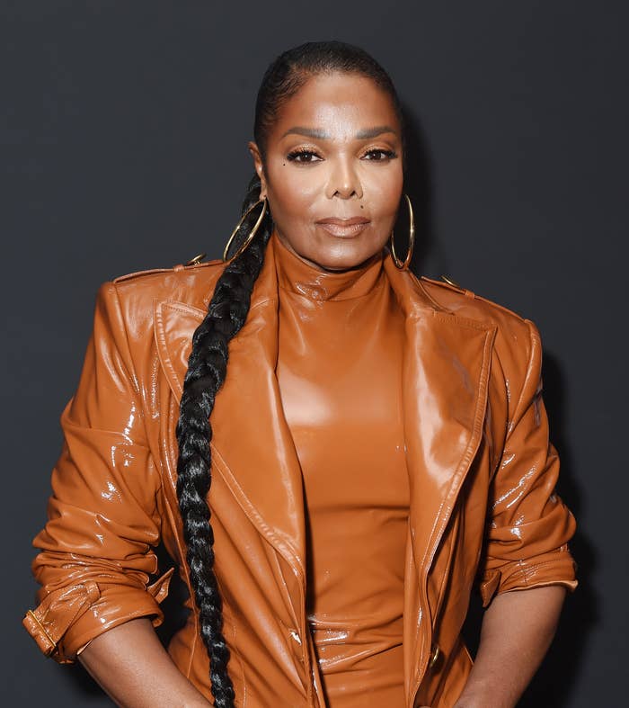 Janet Jackson attending an event, wearing a sleek, high-neck leather outfit and large hoop earrings with a long, braided hairstyle