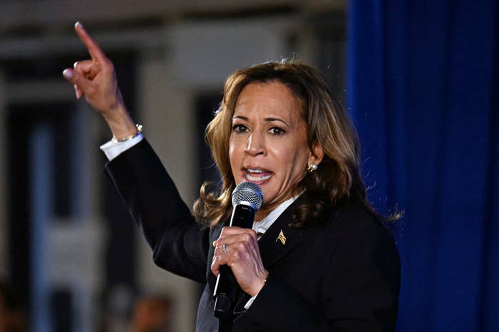 Kamala Harris speaks passionately into a microphone, raising her left hand and pointing upward, wearing a dark suit with a white shirt