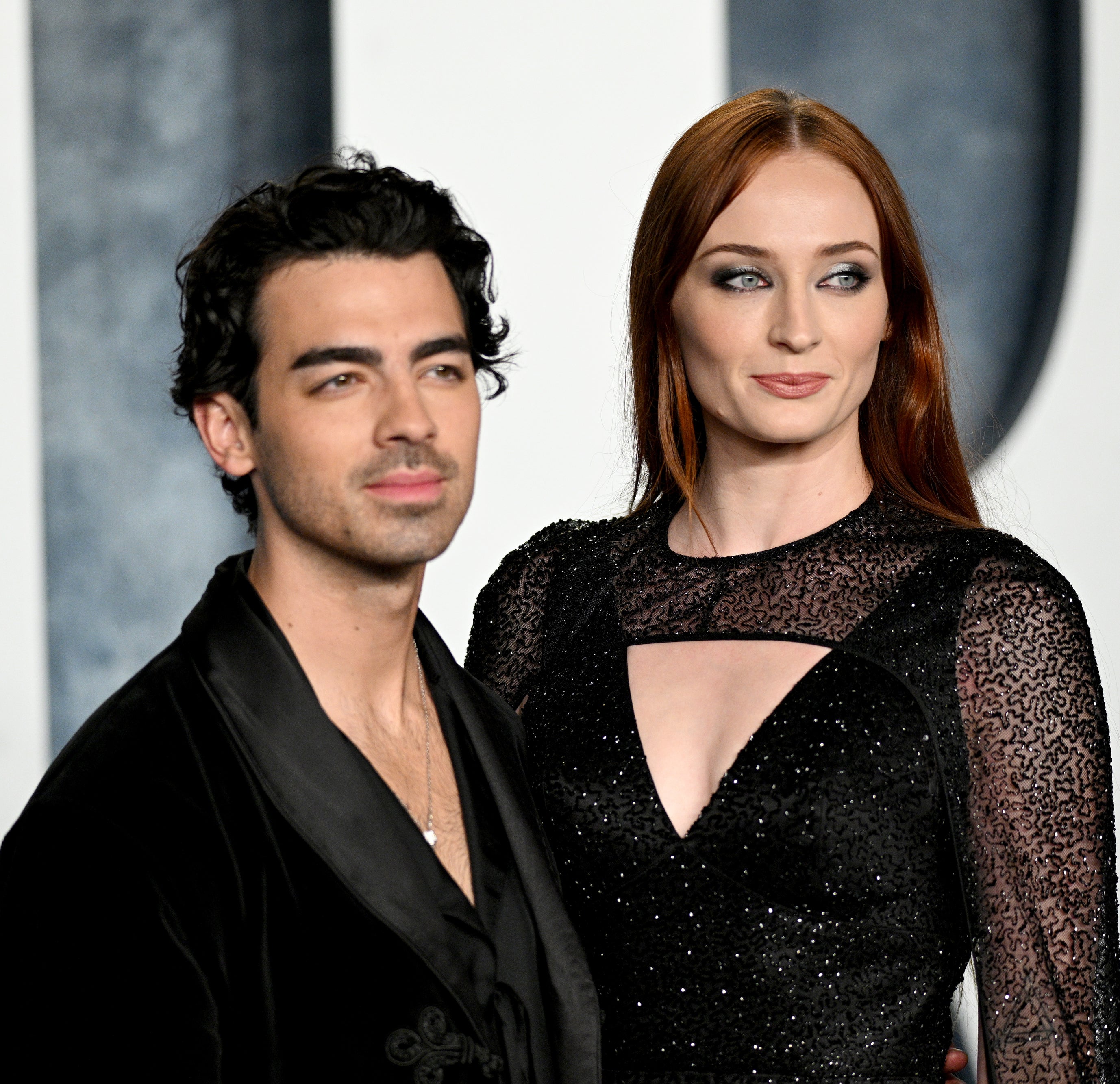 Joe Jonas and Sophie Turner stand together on a red carpet, with Joe in a black outfit and Sophie wearing a black dress with sheer sleeves