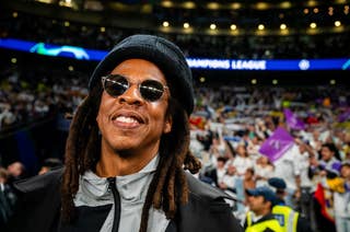 A person with sunglasses and dreadlocks smiles at a sports event, wearing a casual jacket and beanie. Crowd and event signage visible in the background