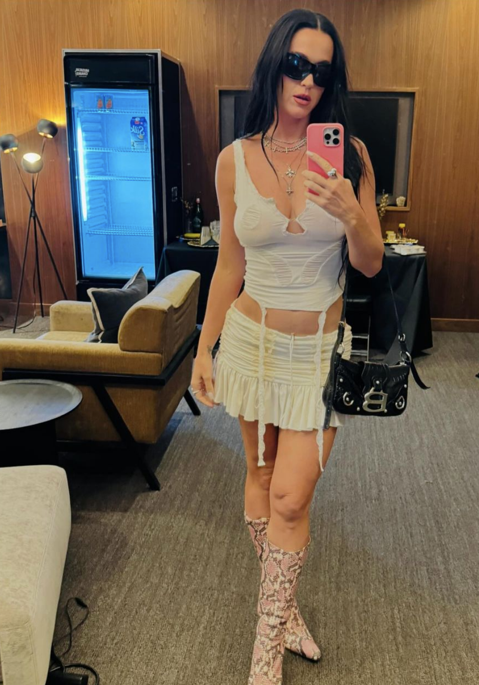 Katy Perry wearing a fitted outfit with a mini skirt and snakeskin boots, holding a phone, posing indoors