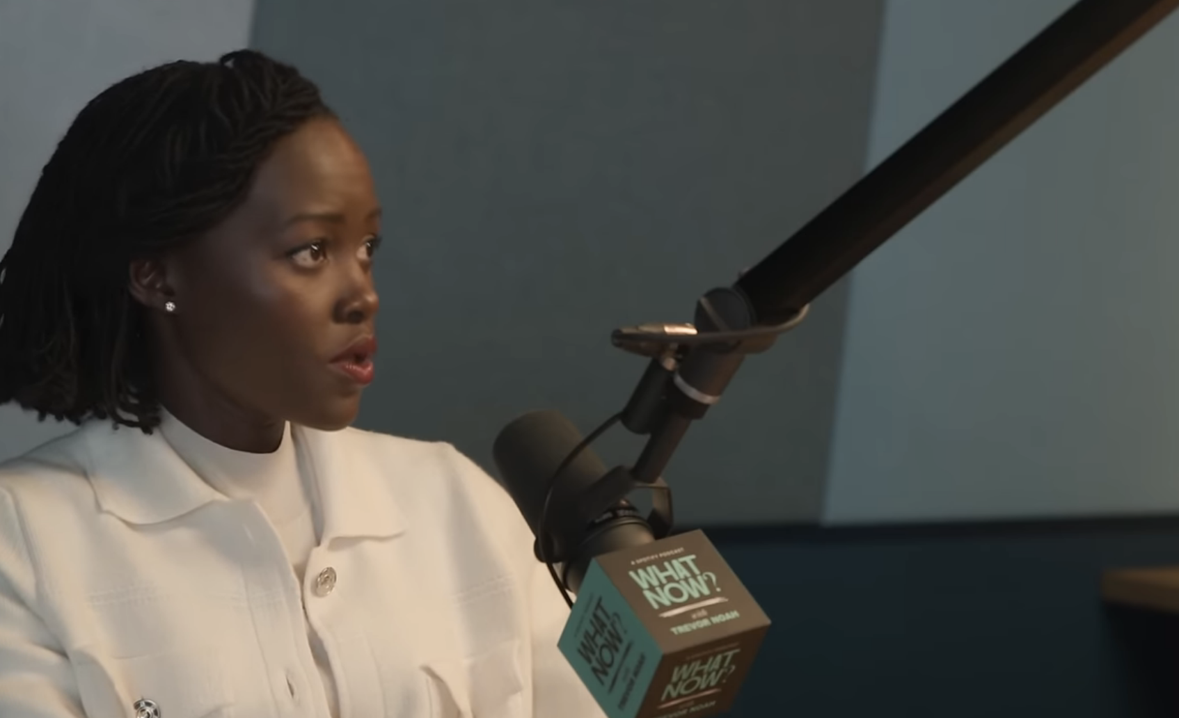 Lupita Nyong&#x27;o in a white outfit talks into a microphone on Trevor Noah&#x27;s podcast, &quot;What Now?&quot; displayed on a YouTube video page